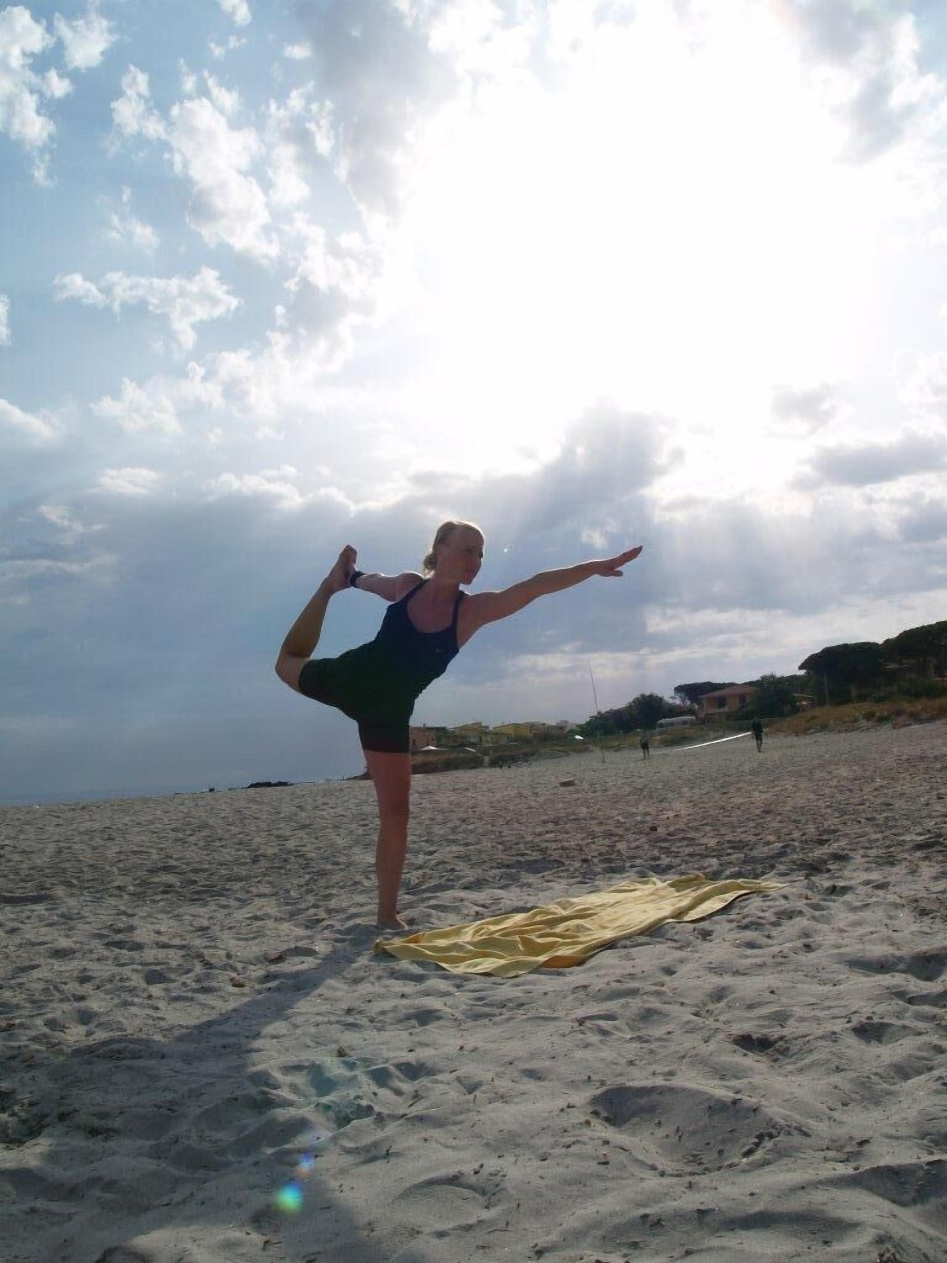 A person performing a yoga pose on a sandy beach with a yellow towel and buildings in the background.