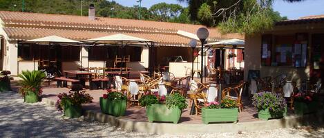 An outdoor café with tables and chairs, surrounded by potted plants and a tiled roof.