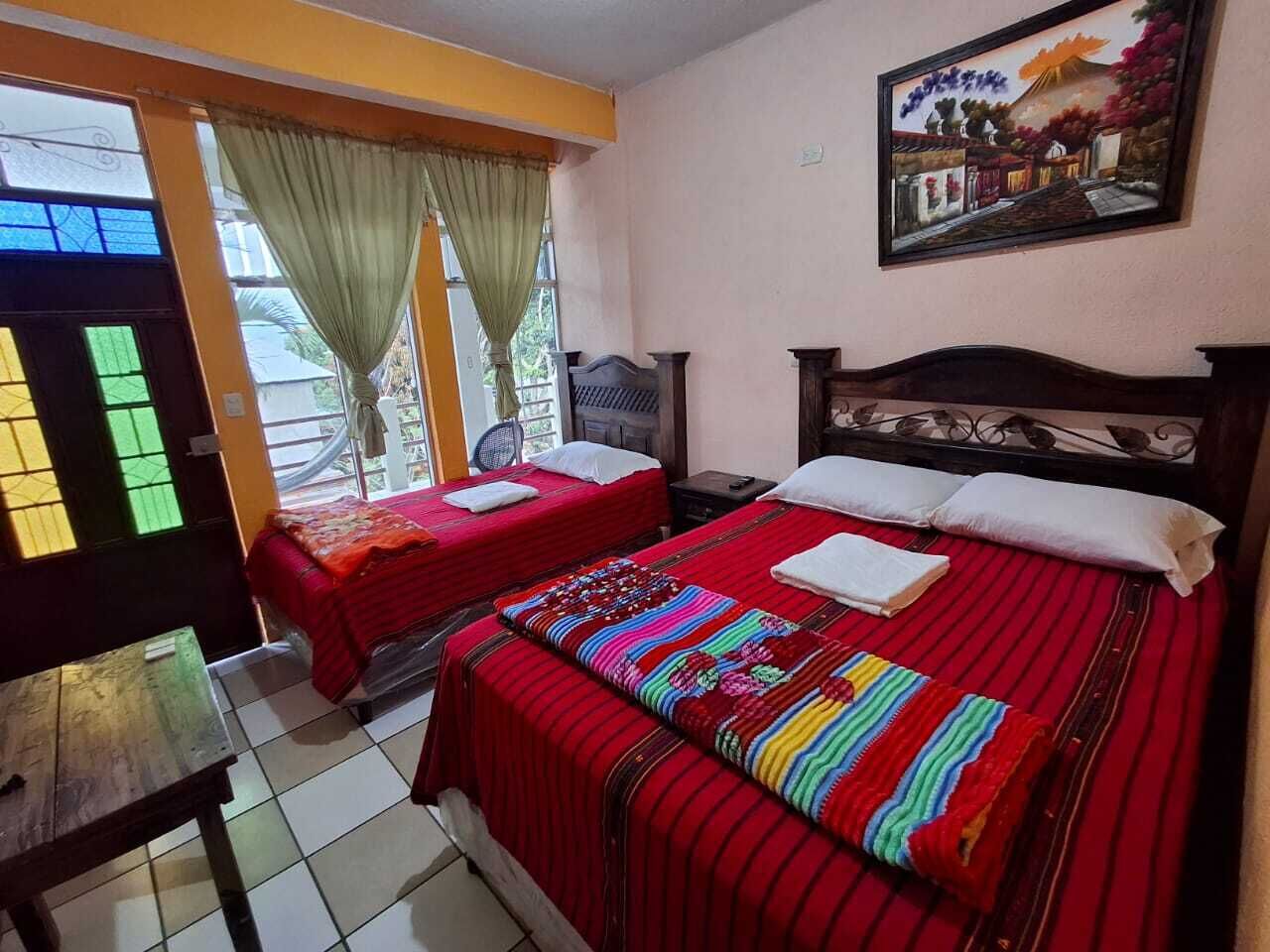 A hotel room with two beds, a wooden table, a window with curtains, and a painting on the wall.