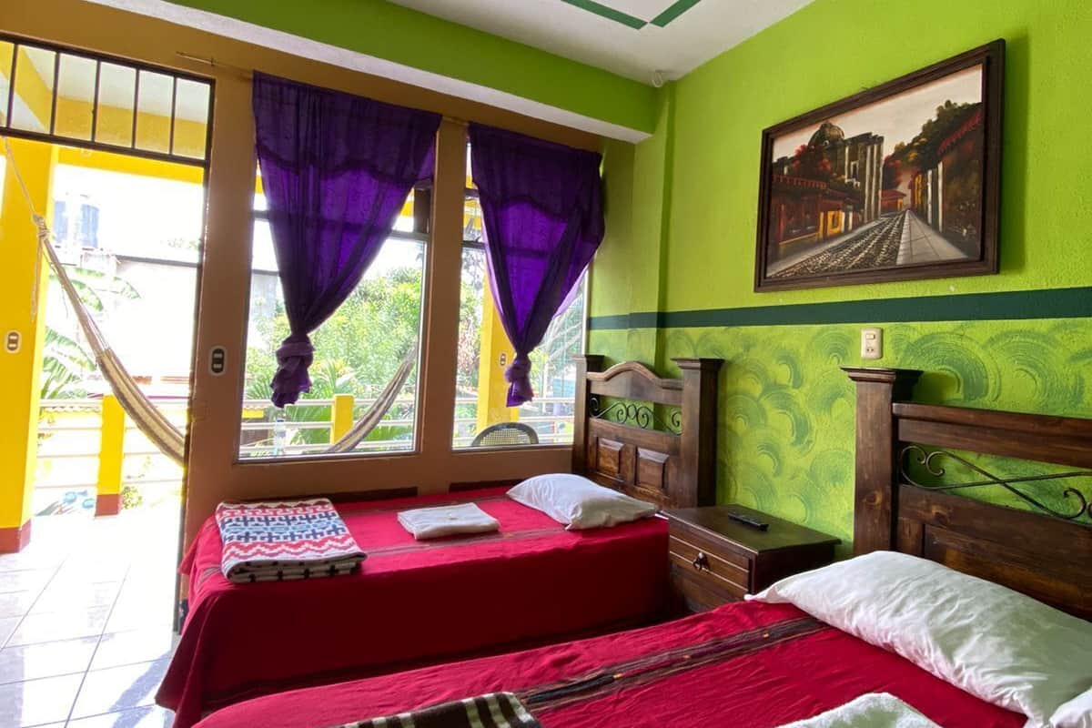 Une chambre avec deux lits, une fenêtre avec des rideaux violets, un tableau au mur et un ventilateur de plafond.
