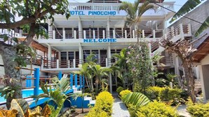 Entrada de un hotel con un cartel de 'Welcome', zona de piscina y jardín con diversas plantas.