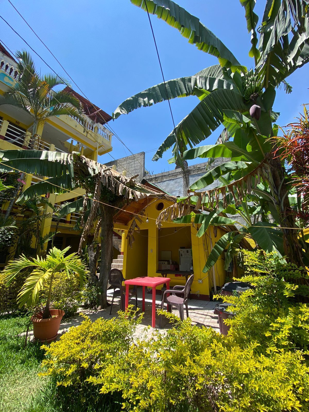 Um edifício amarelo com uma mesa e cadeiras vermelhas no pátio, rodeado por vegetação exuberante e bananeiras.