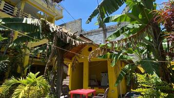 Um edifício amarelo com uma mesa e cadeiras vermelhas no pátio, rodeado por vegetação exuberante e bananeiras.