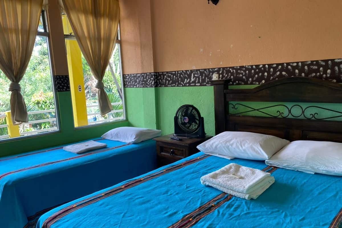 Une chambre avec deux lits, chacun doté de draps bleus et d’oreillers blancs, d’une tête de lit en bois, d’une petite table de chevet avec un ventilateur et d’une fenêtre avec des rideaux.