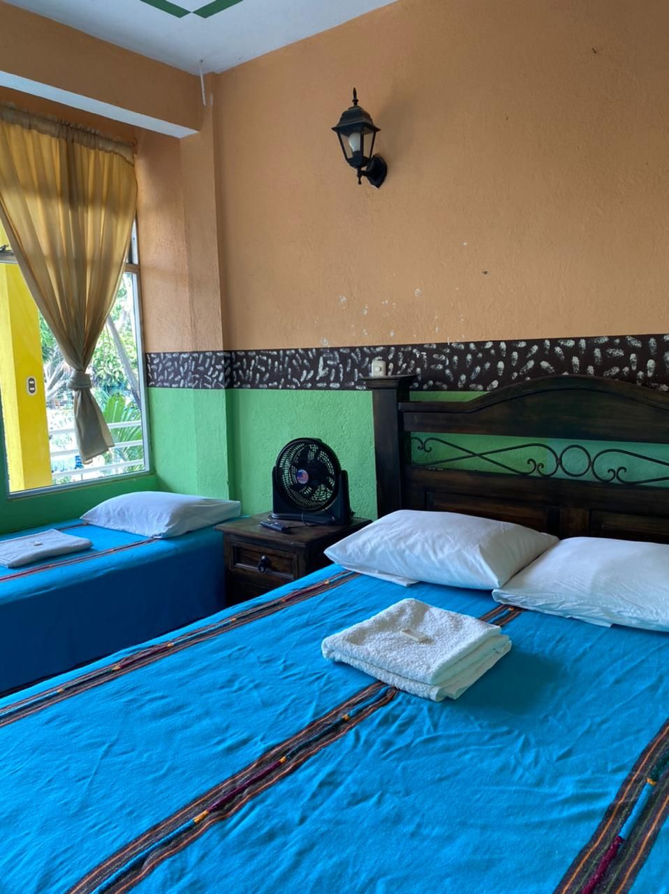 A room with two beds, a nightstand with a fan, a wall-mounted lamp, and a window with curtains.
