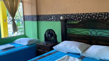 A room with two beds, a nightstand with a fan, a wall-mounted lamp, and a window with curtains.