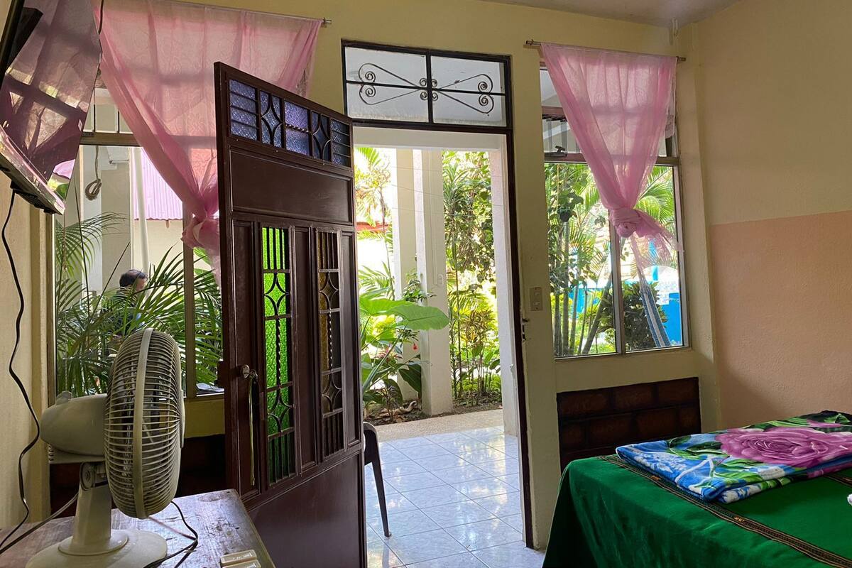 Une chambre avec un lit, un ventilateur et une vue sur un jardin, visible par une porte.
