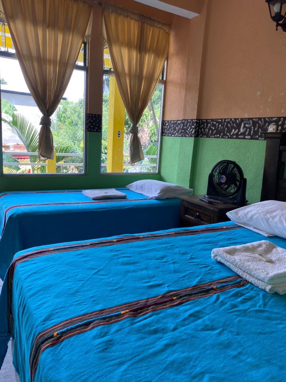 Une chambre avec deux lits, chacun agrémenté de draps bleus et d’oreillers blancs, une petite table avec un ventilateur et une vue sur la verdure environnante.