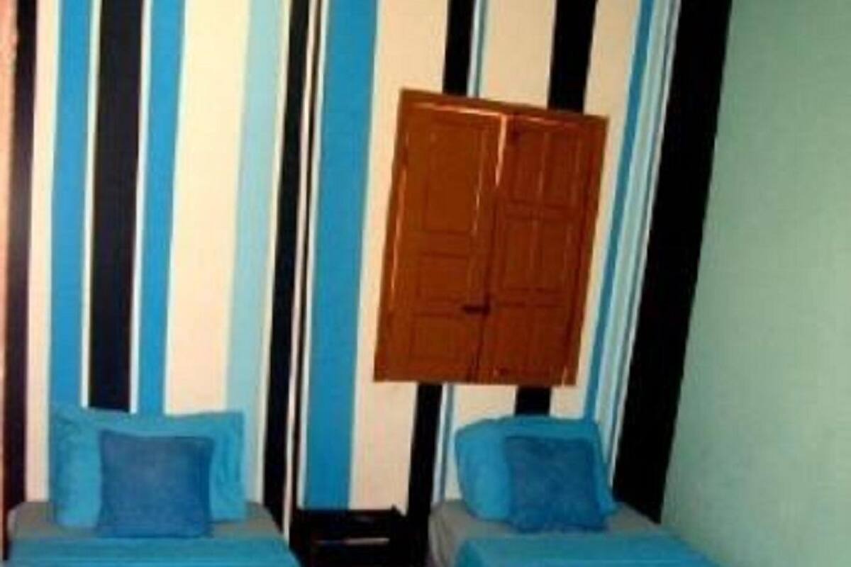Ein Zimmer mit zwei Einzelbetten, jeweils mit einer blauen Bettdecke und einem weißen Handtuch. Es gibt gestreifte Vorhänge und eine Holztür.