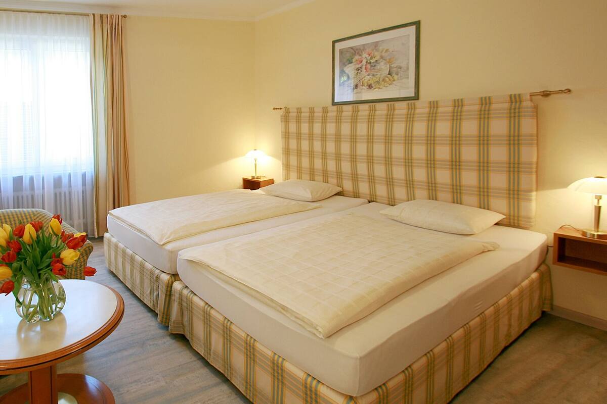Una habitación de hotel con dos camas, una mesita de noche con una lámpara, un jarrón de tulipanes y un cuadro en la pared.