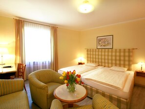 Quarto de hotel com cama, duas mesas de cabeceira, uma cadeira, uma mesa redonda com um vaso de flores e uma janela com cortinas.