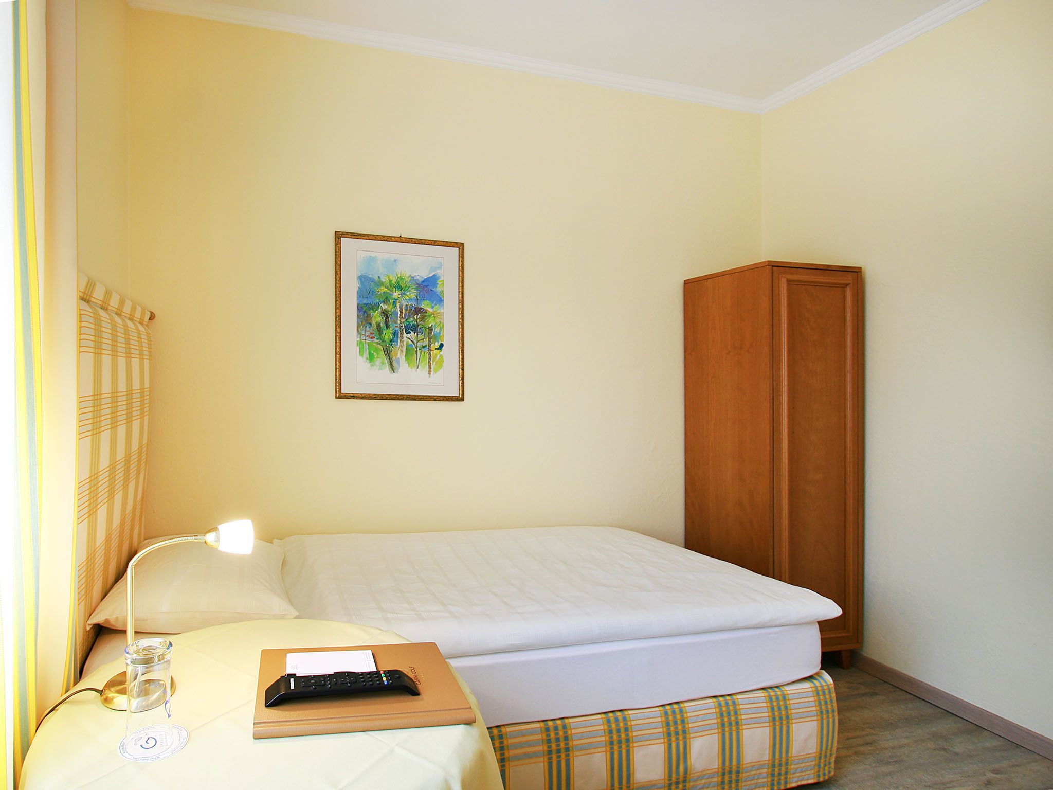 Un dormitorio pequeño y ordenado con una cama individual, una mesita de noche, una lámpara, un control remoto y un cuadro en la pared.