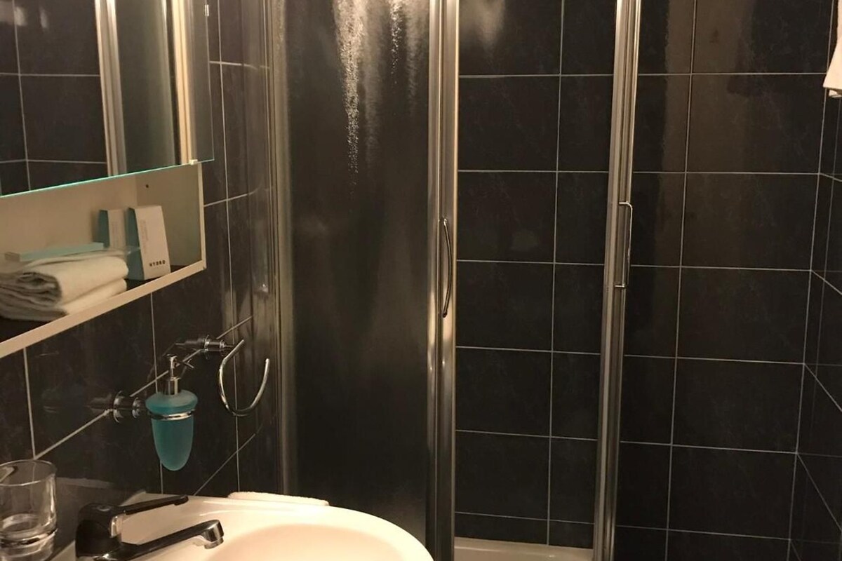 Casa de banho com chuveiro em cabine de vidro, lavatório branco e espelho sobre o lavatório.