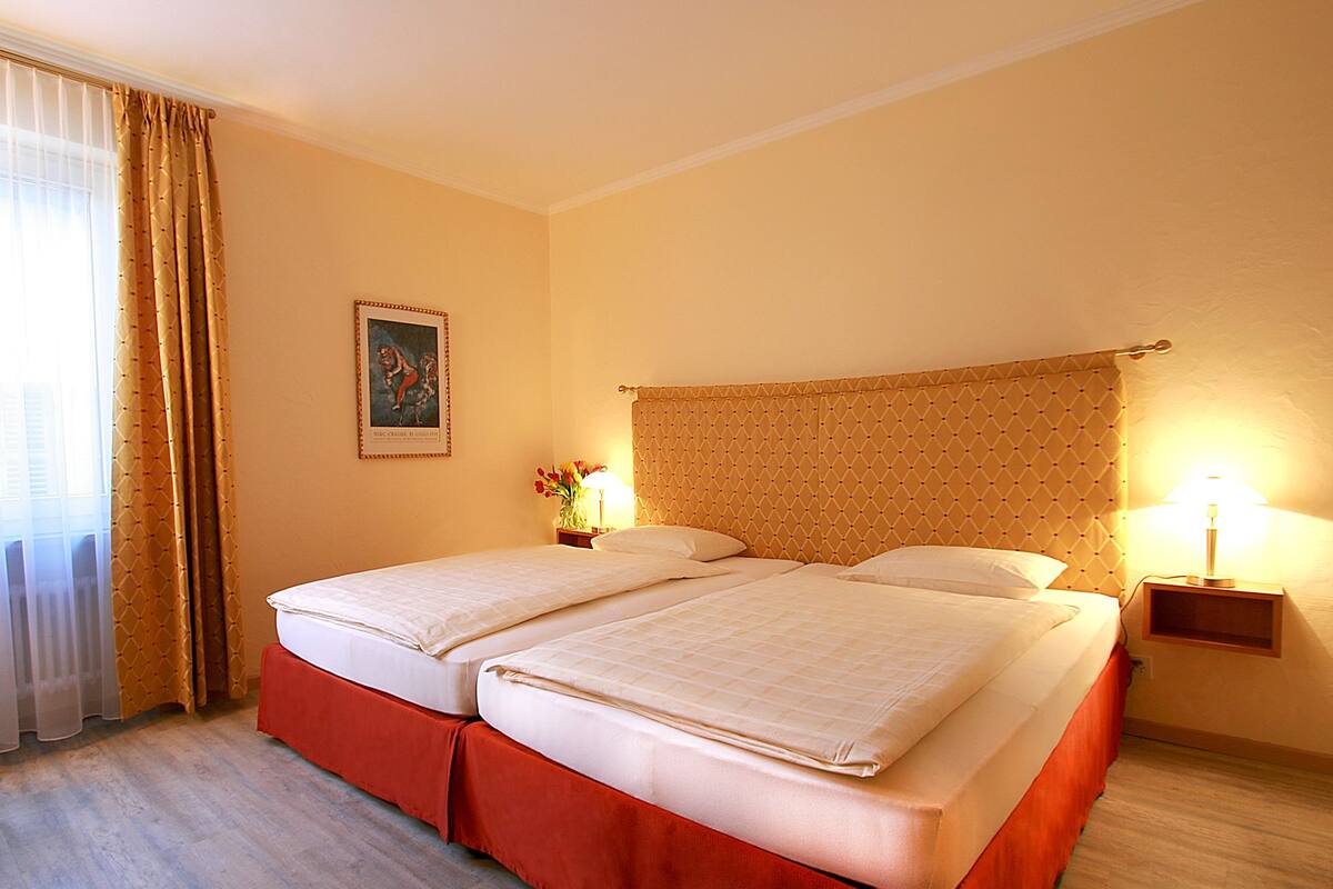 Una cama doble con falda roja, una mesita de noche con lámpara y un cuadro en la pared.