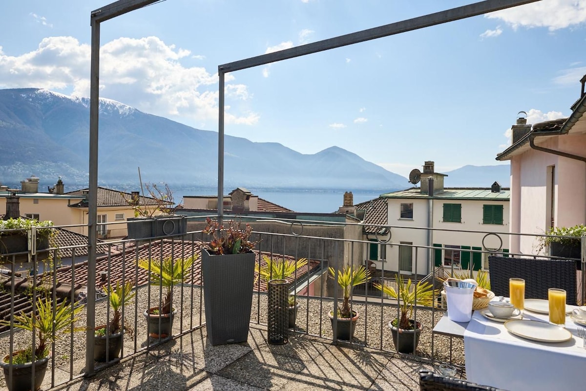 A terrace with potted plants, a table set for breakfast, and a view of the sea and mountains.