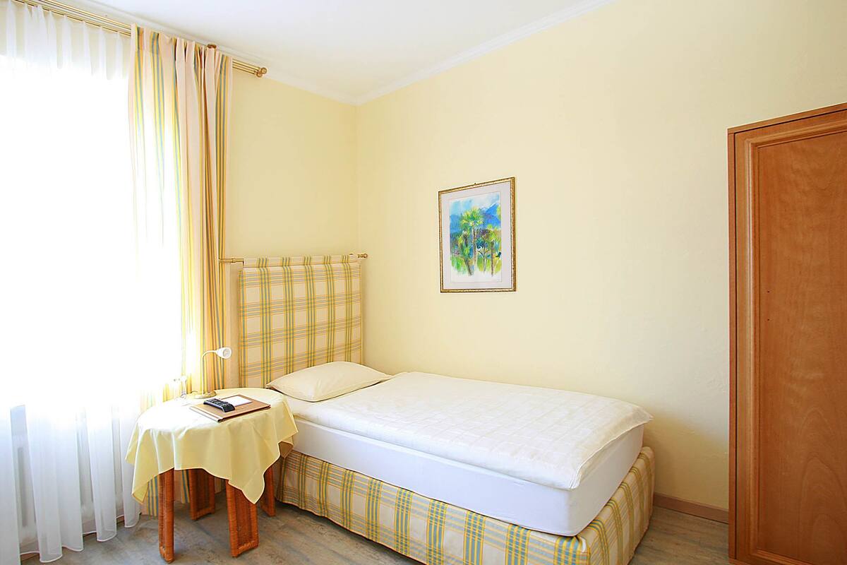 Una habitación pequeña con cama individual, un armario de madera, una mesita de noche con lámpara y un cuadro en la pared.