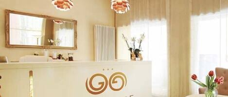 A reception area with a white counter, a large mirror, and a logo for 'Garn Golf'. There are two pendant lights above the counter, and a vase with flowers on the right side.
