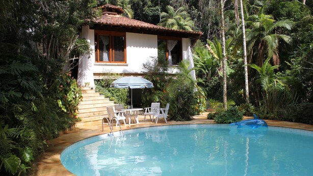 A swimming pool with a dolphin float, surrounded by lush greenery and a house with a tiled roof. - Refugio Em Teresopolis (Teresópolis)