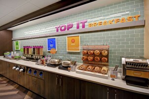 Una estación de desayuno con un letrero que dice "TOP IT BREAKFAST" y diversas opciones de comida como waffles, huevos y pasteles.