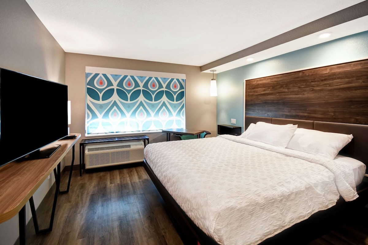 A hotel room with a bed, a TV, a heater, a window with a patterned view, and a wooden headboard.