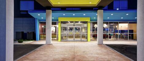 A modern building entrance with a glass door, a yellow awning, and a paved area with plants.