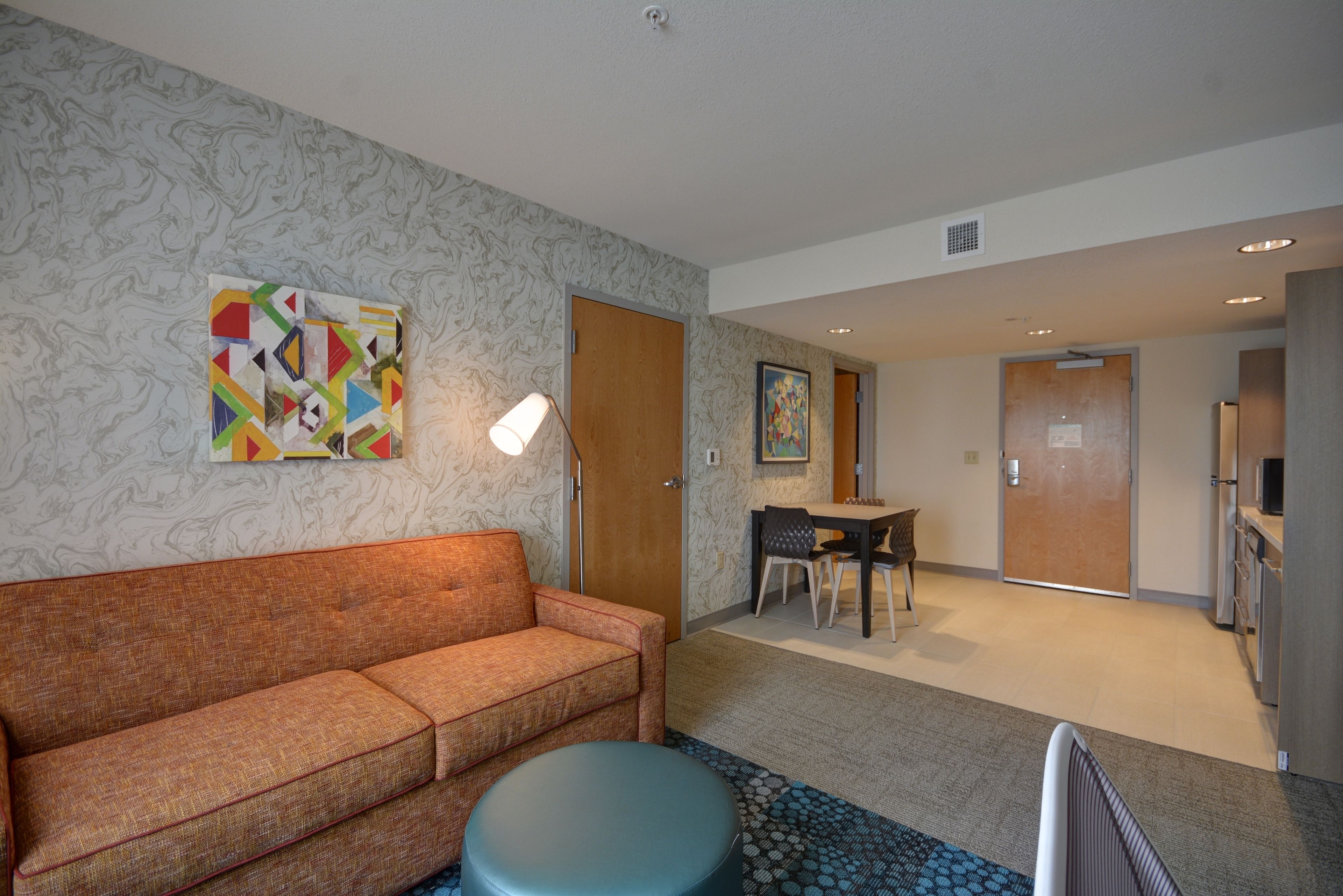 A hotel room with a sofa, a round ottoman, a dining area with a table and chairs, and a kitchenette.