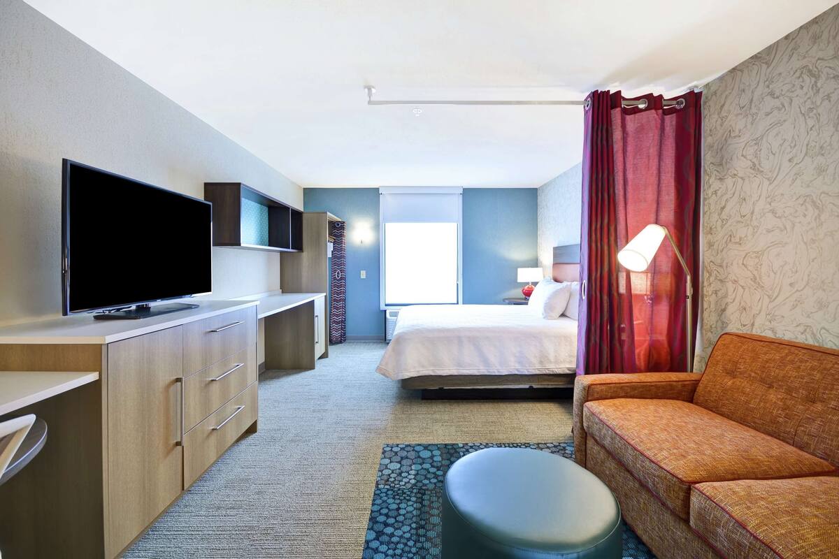 Ett hotellrum med en platt-TV, en säng, en soffa, ett skrivbord och ett fönster med gardiner.