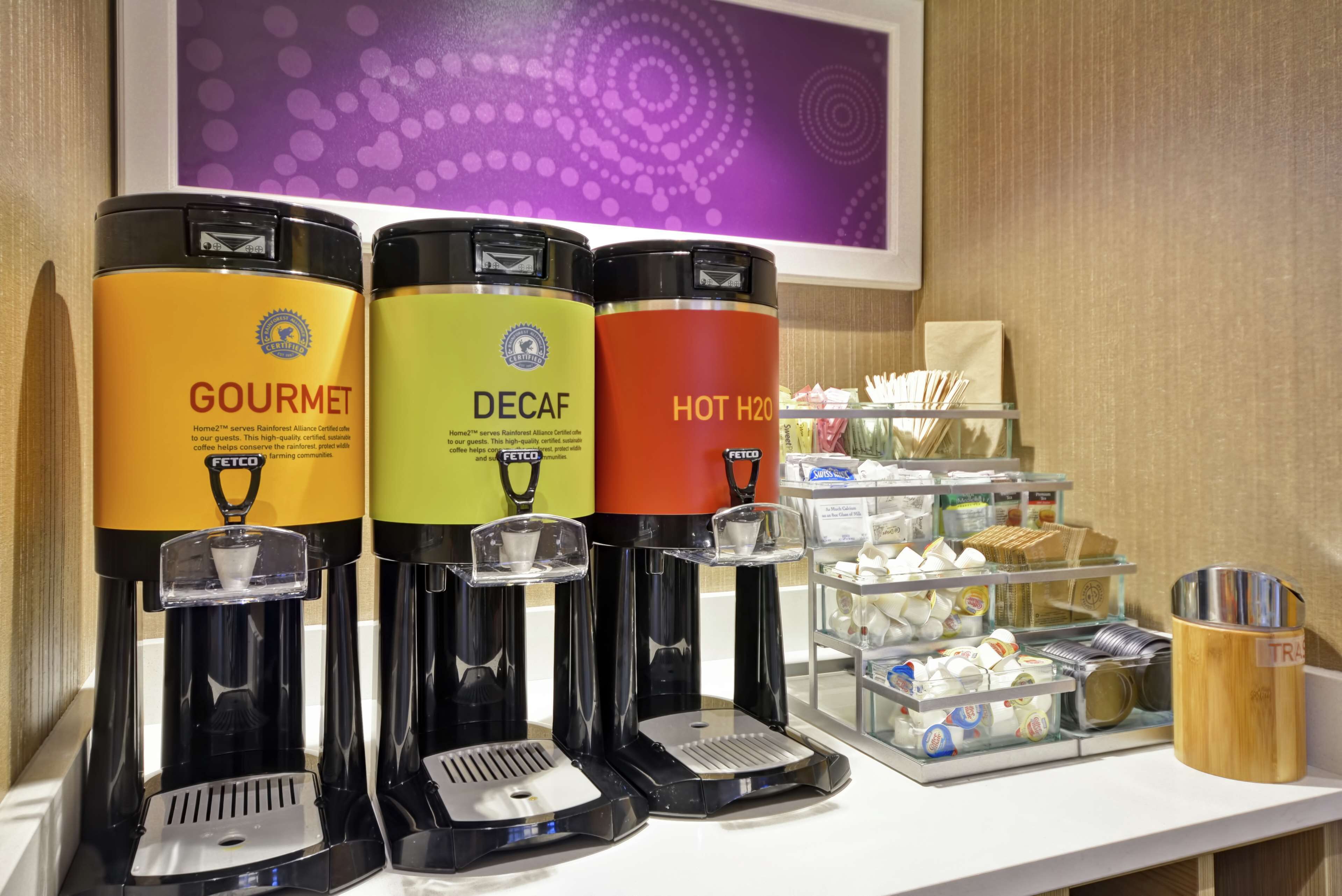 Three beverage dispensers labeled 'GOURMET,' 'DECAF,' and 'HOT H2O' on a counter with a tray of snacks and a trash can.