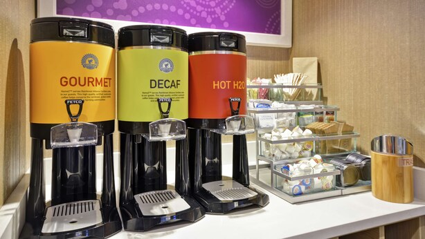 Three beverage dispensers labeled 'GOURMET,' 'DECAF,' and 'HOT H2O' on a counter with a tray of snacks and a trash can. - Home2 Suites by Hilton Portland Airport (South Portland)