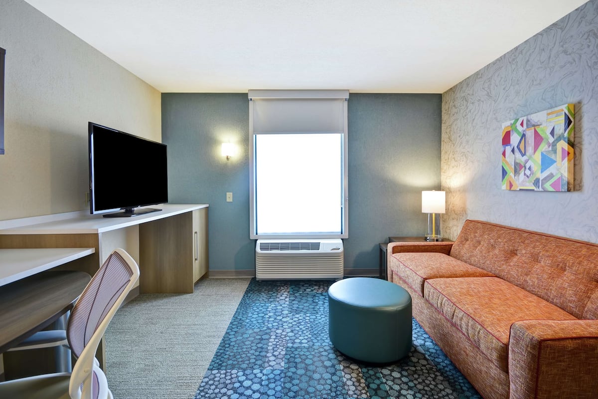 Ett hotellrum med en platt-TV, en soffa, en pall, ett skrivbord och ett fönster med utsikt.