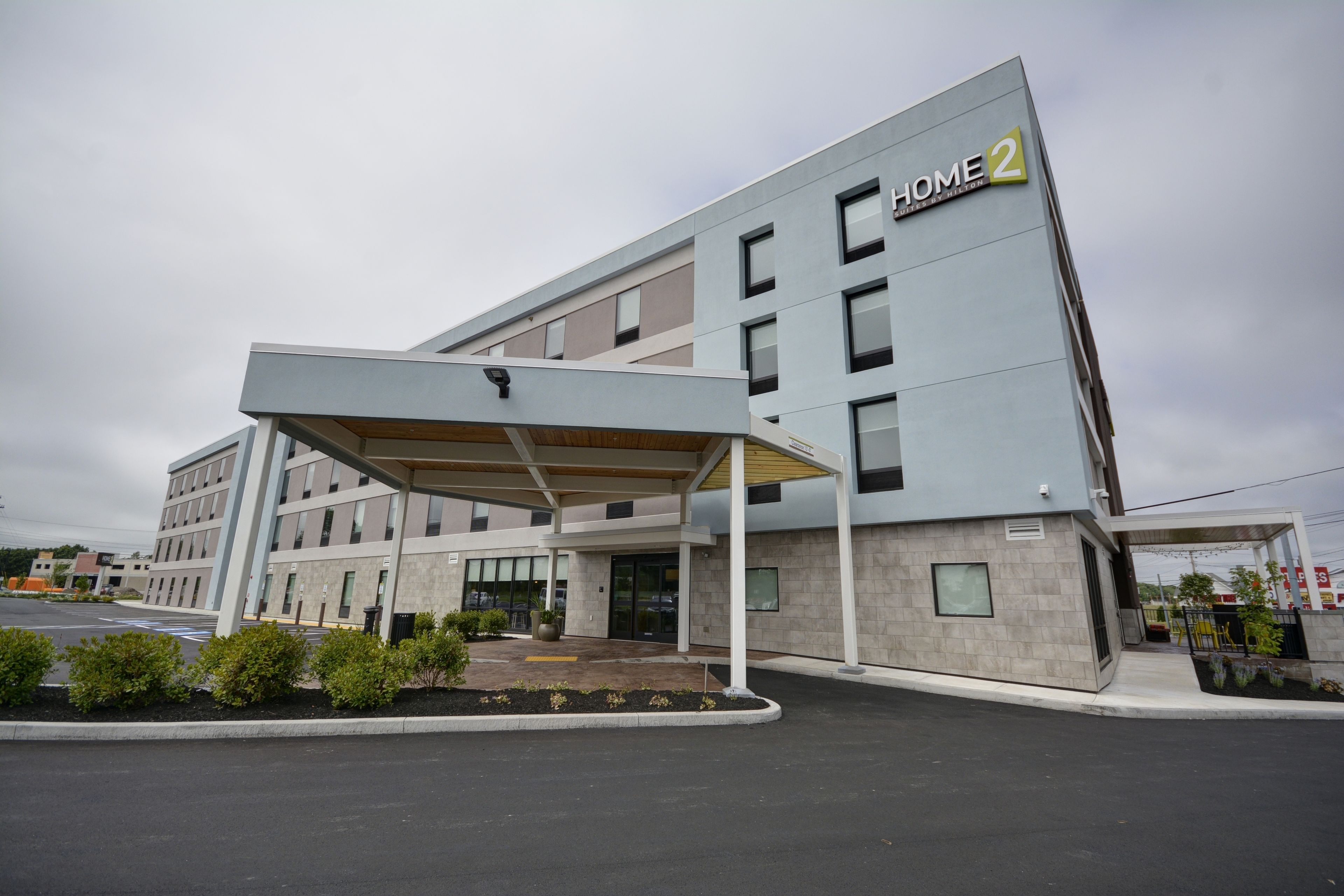 A modern hotel building with a covered entrance, multiple floors, and a sign indicating it is "Home 2".