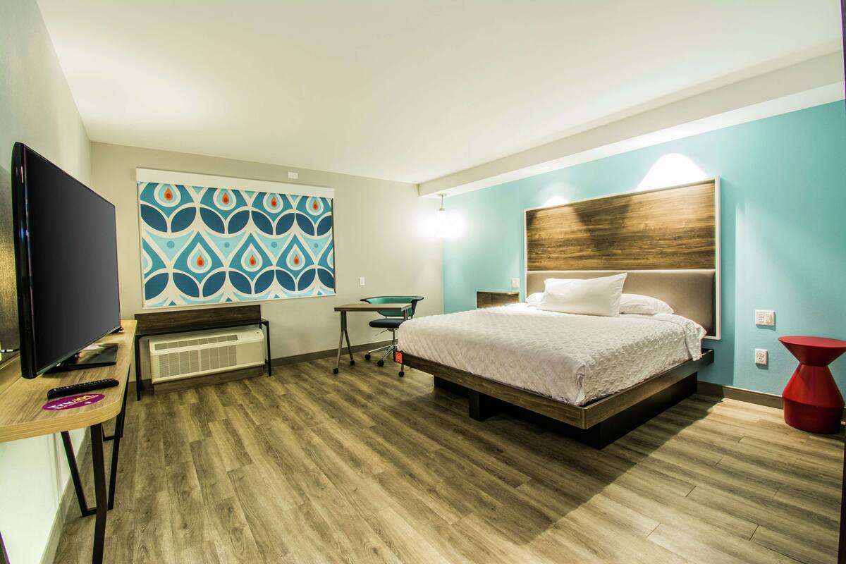 A hotel room with a bed, a desk with a TV, a chair, and a wall with a geometric pattern.