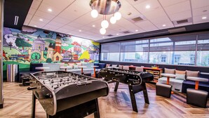 Un espacio interior moderno con dos mesas de fútbol de mesa, un mural que representa un paisaje urbano y una zona de estar con mesas y sofás.