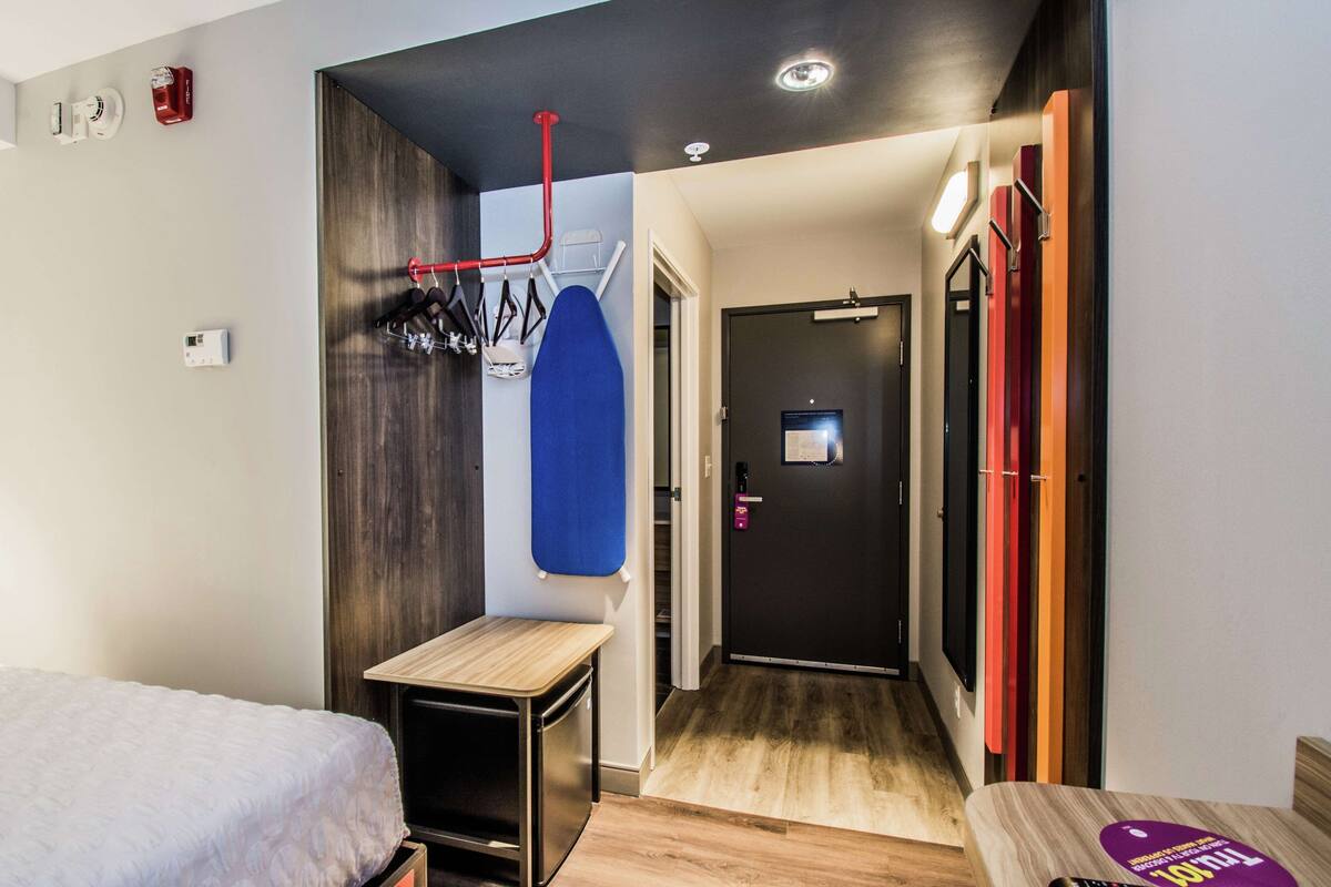 A compact hotel room with a bed, a small table, a blue towel, a coat rack, and a door leading to a hallway with a mirror and a thermostat.