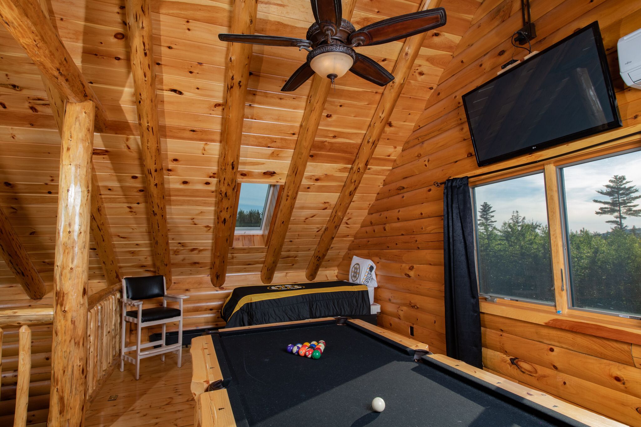 A wooden cabin with a pool table, a ceiling fan, a television, and a window with a view of trees.