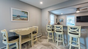 Un coin repas avec une table et quatre chaises, un tableau représentant une scène de plage, et un espace cuisine avec une fenêtre et un ventilateur de plafond.