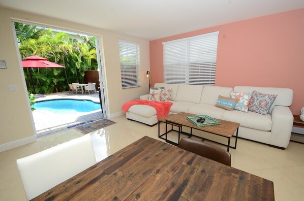 A living room with a white sofa, a wooden coffee table, and a dining table. There is a pool visible through the glass door.