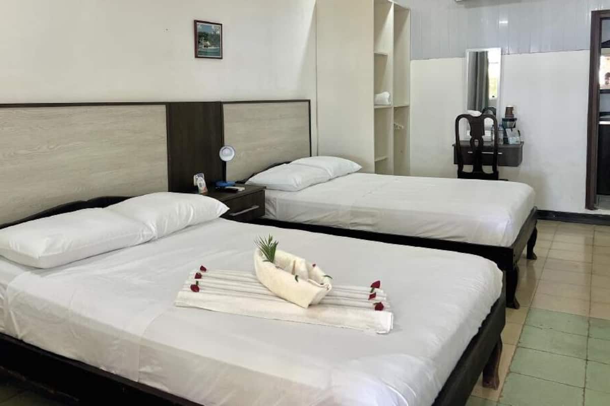 A hotel room with two beds, a bedside table with a lamp, a mirror, and a small shelf with toiletries.