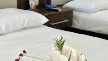 Una cama bien hecha con sábanas blancas, una toalla doblada con un ramita de verdor y algunos pétalos de rosa sobre la cama.