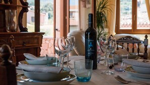 Una mesa de comedor dispuesta con platos, tazones y copas de vino, con una botella de vino y una planta al fondo.