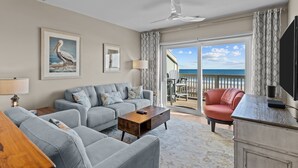 Een woonkamer met een grijze bank, een houten salontafel en een rode fauteuil. Er is een groot raam met uitzicht op het strand en de oceaan.