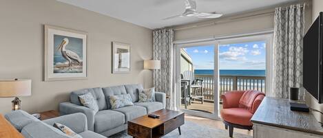 A living room with a grey sofa, a wooden coffee table, and a red armchair. There is a large window with a view of the beach and ocean.