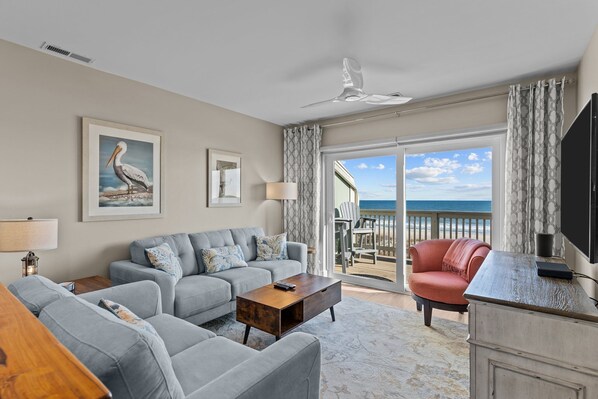 A living room with a grey sofa, a wooden coffee table, and a red armchair. There is a large window with a view of the beach and ocean.