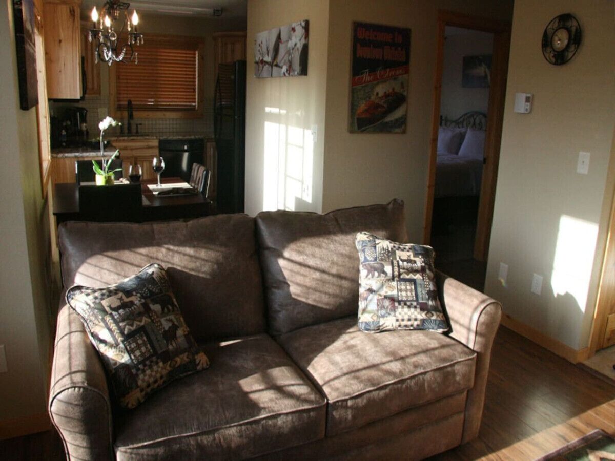 A living room with a brown sofa, two patterned throw pillows, a chandelier, and a kitchen in the background.