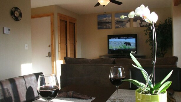A dining area with a table set for two, two wine glasses, a potted orchid, and a television displaying Glacier National Park.
