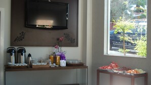 A breakfast buffet setup with a TV above, a window with a view of a parked car, and a small table with fruits and snacks. - Hotel Recanto dos Ipês (Frutal)