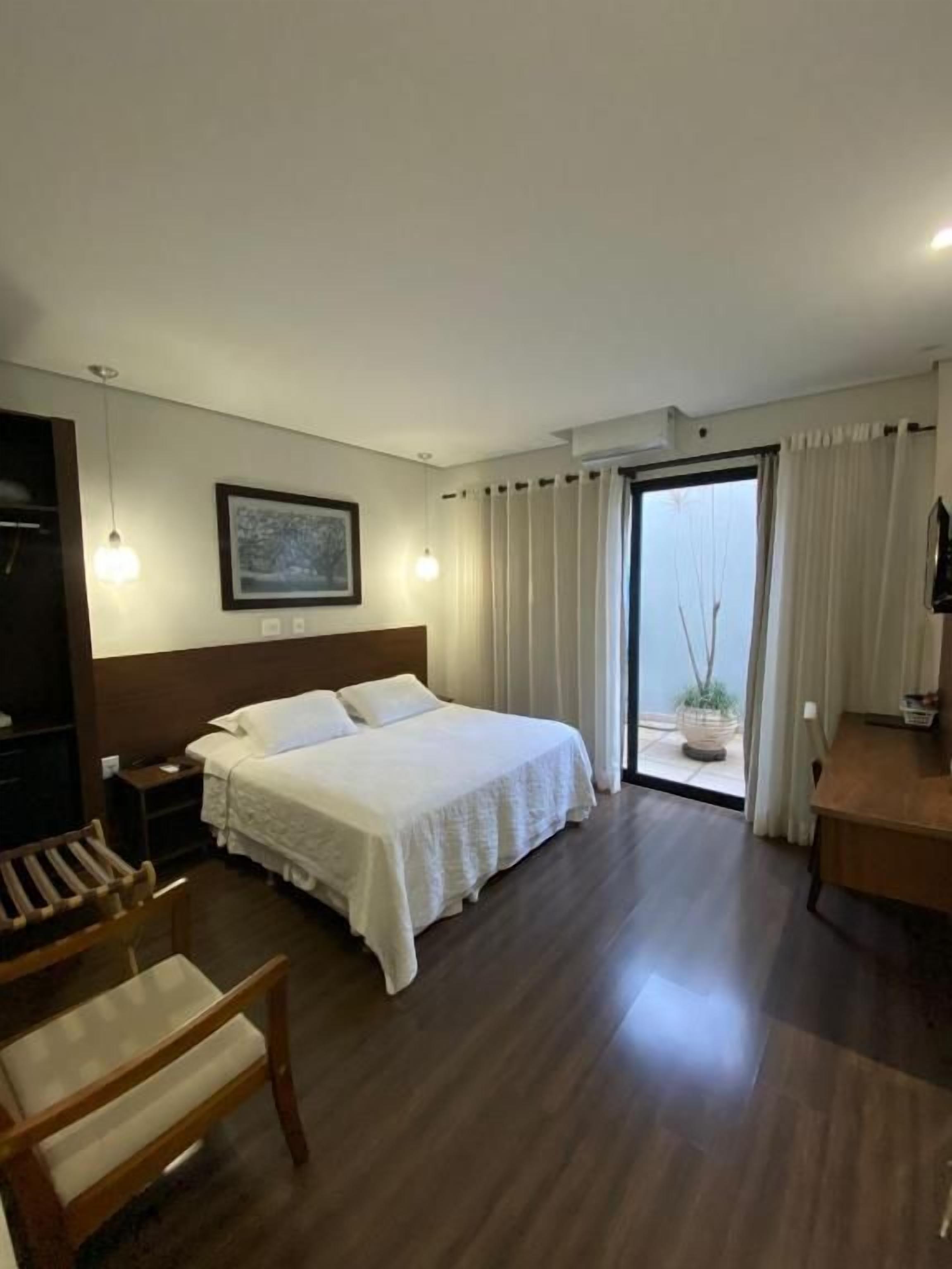 A hotel room with a large bed, a wooden chair, a desk, and a view of the outdoors through a sliding door.