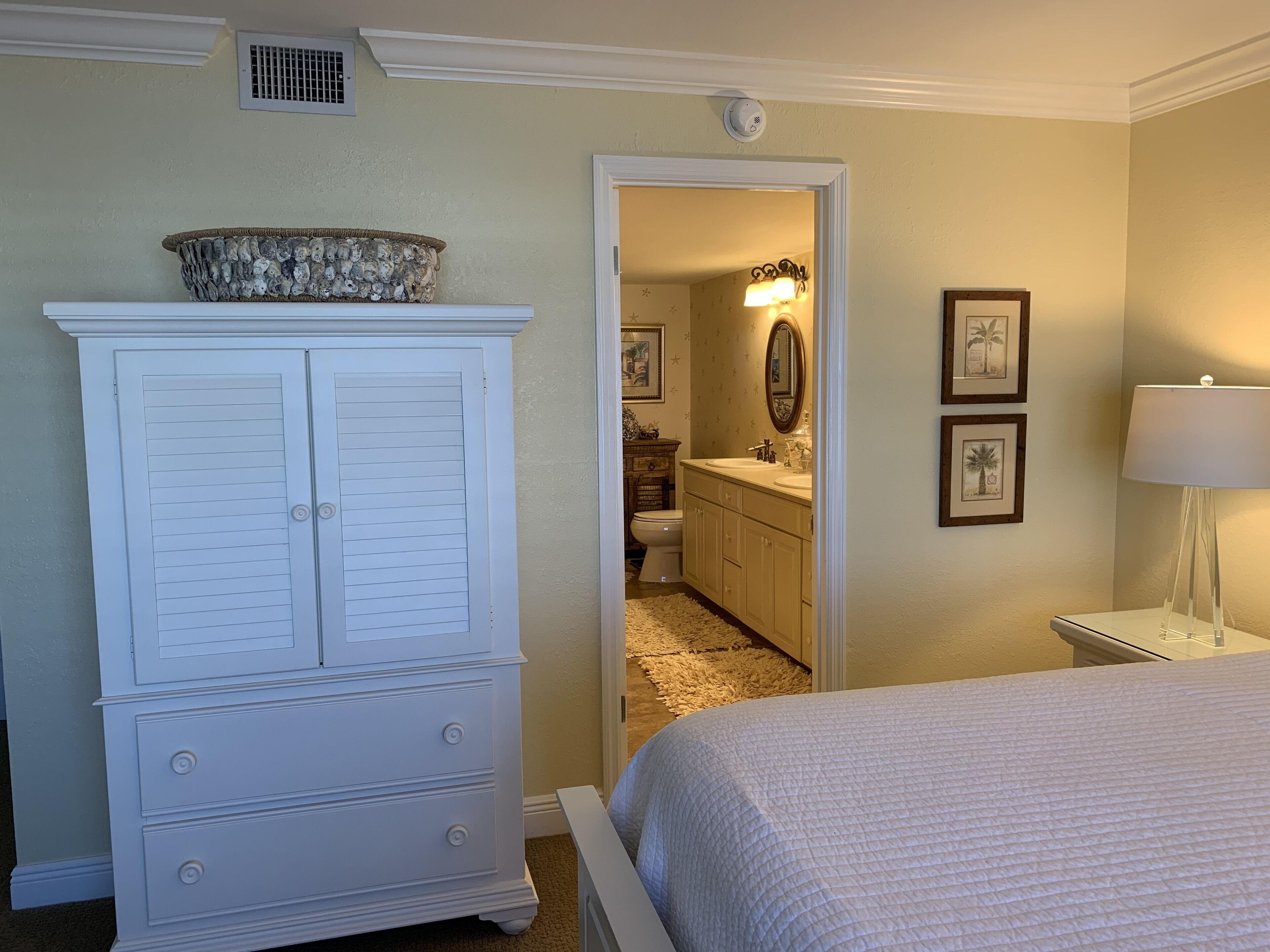 A bedroom with a bed, a dresser, a bathroom visible through an open door, and a nightstand with a lamp.
