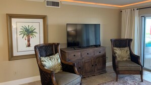 A living room with two wicker chairs, a wooden entertainment center, a framed picture of a palm tree, and a large flat-screen TV. - Ground floor with King master, 2 twins 2nd bedroom Bunk beds in 3rd bedroom (twins) twin/full combo. (New Smyrna Beach)