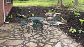 A patio with stone flooring and green metal chairs, surrounded by a garden with various plants and a stone wall. - Watkins Glen lake view rental (Watkins Glen)